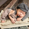 A newborn baby sleeping wearing a gray knit romper and newsboy cap, resting on a light-colored fabric.