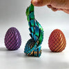 Eine Hand hält ein verdrehtes Drachen-Ei-Fidget-Spielzeug mit irisierenden grün- und blauen Schuppen, neben zwei solide lila 