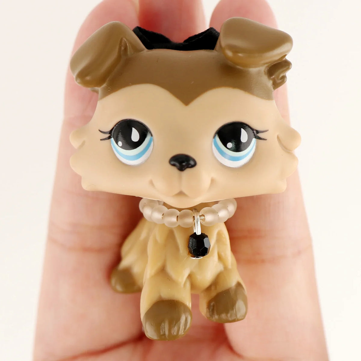 A small, light brown miniature pet animal figure with large blue eyes and a black collar is held in a person's hand.