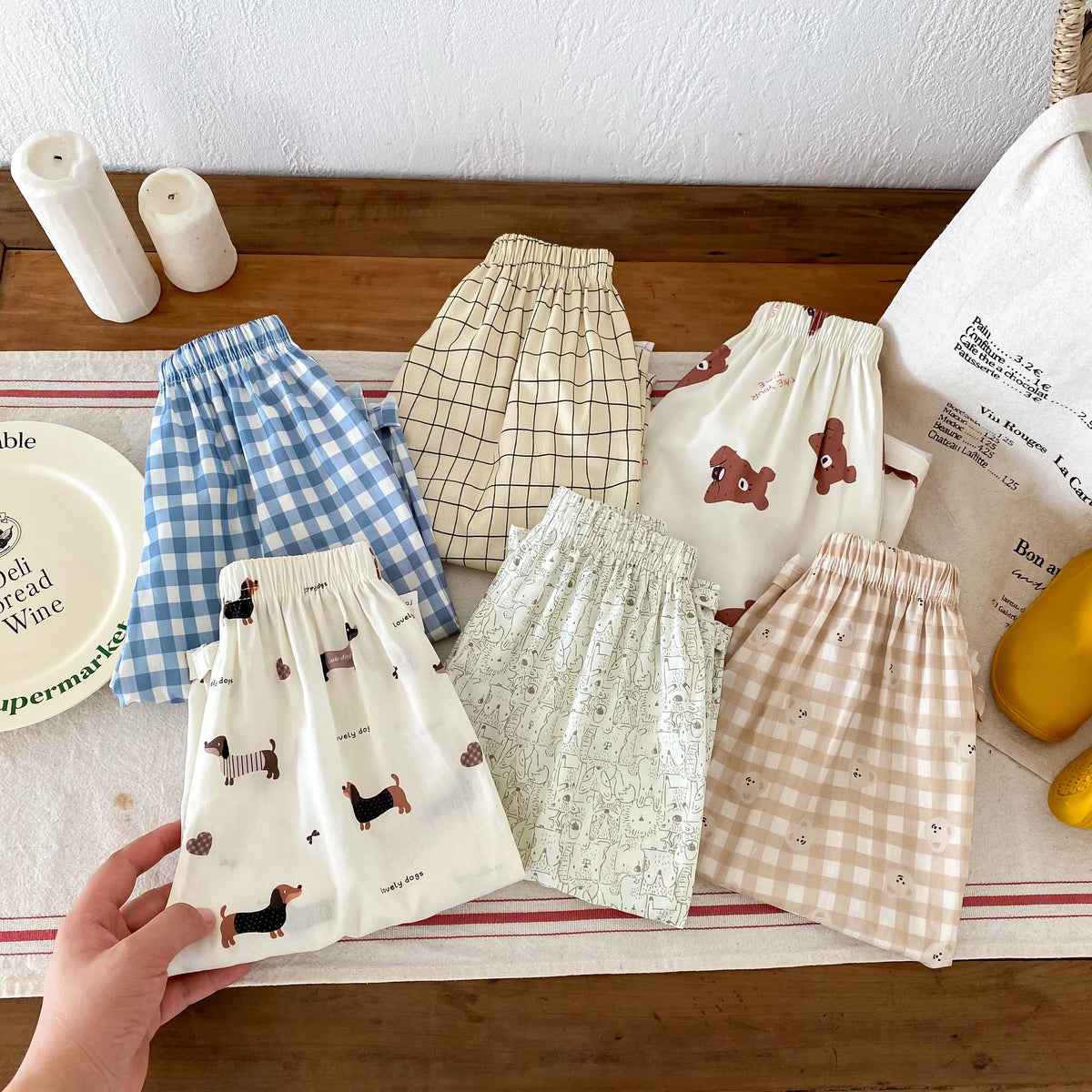 A collection of kids casual pants in various plaid and patterned fabrics laid out on a wooden surface.