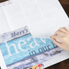 A hand holds a sheet of double-sided non-stick release paper for diamond painting featuring text and colorful designs.