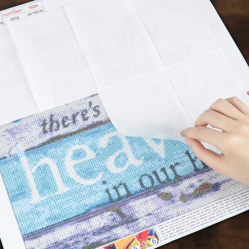 A hand holds a sheet of double-sided non-stick release paper for diamond painting featuring text and colorful designs.