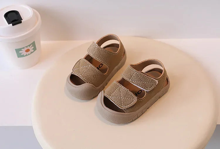 A pair of tan suede toddler soft-soled sandals for boys and girls resting on a light colored surface.