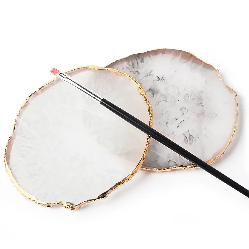 A round, translucent white resin palette with a gold trim and a thin brush resting on it.