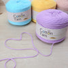 A collection of skeins of soft cotton yarn in blue, yellow, and purple is displayed on a light wooden surface.