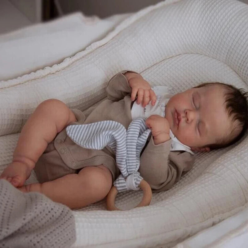 A newborn baby doll with rooted hair is lying in a white fabric cradle.