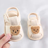 A pair of light beige soft sole sandals for a baby, featuring a cute bear face design.