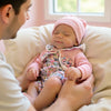 A newborn baby wearing a pink knitted hat and floral outfit is being held by an adult in a soft, light-colored setting.