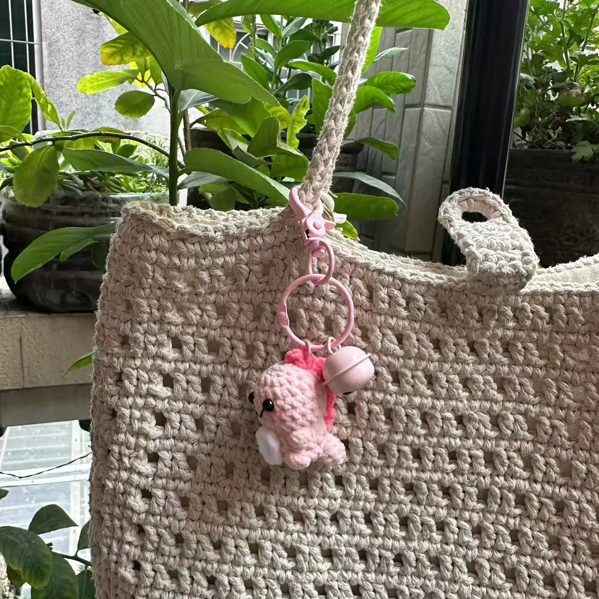 A handmade crochet pink dinosaur keychain hanging from a beige woven bag