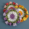 A circular arrangement of hand-crocheted floral coasters in various shades of pink, purple, yellow, and green.