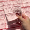 A hand holds a white strawberry milk squishy stress relief toy against a pink grid background.