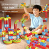 A smiling boy is building a colorful magnetic train track set with various shapes on a wooden floor.