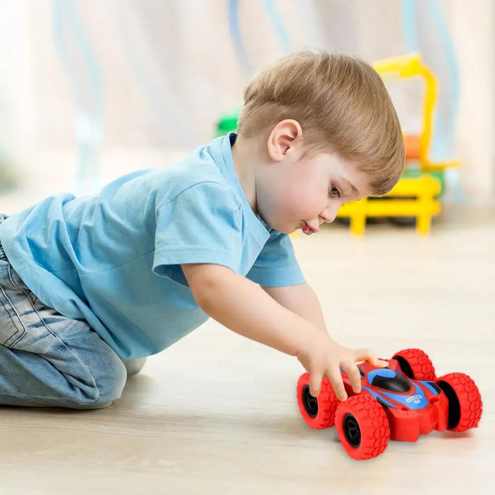 A young boy is playing with a red and blue four-wheel drive stunt car on a light wooden floor.