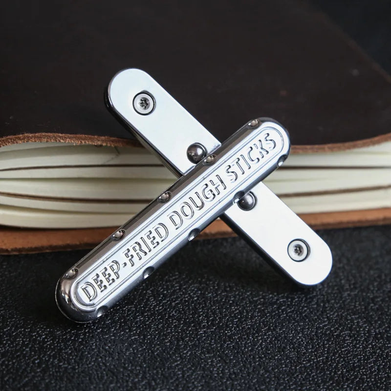 A silver metal fidget slider labeled DEEP-FRIED DOUGH STICKS is shown crossed on a dark surface next to a stack of books.