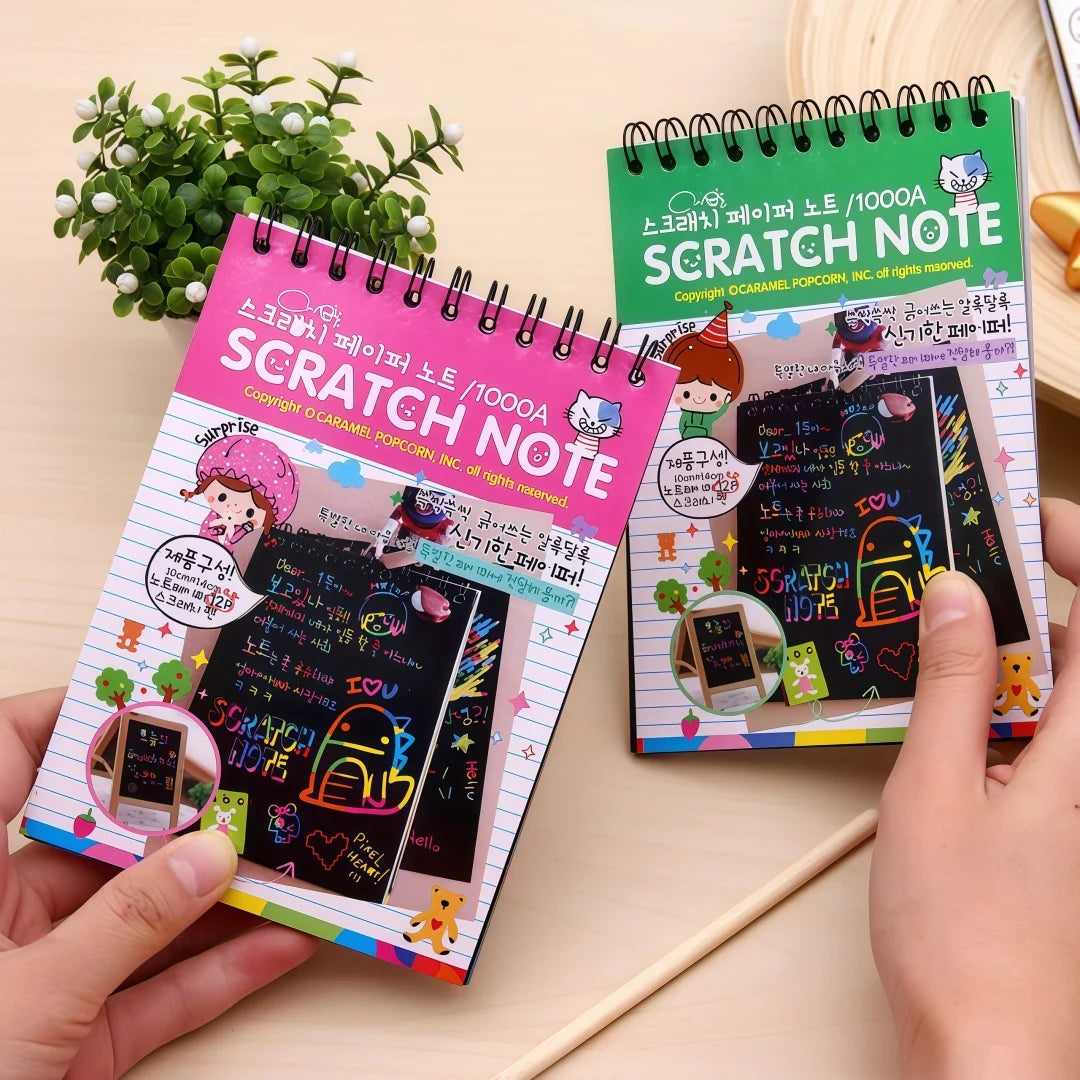A hand holds a pink Scratch Note notebook featuring colorful scratch art designs and a green spiral notebook.