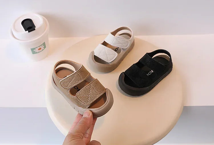 A pair of beige soft-soled sandals for toddlers is shown on a light surface.