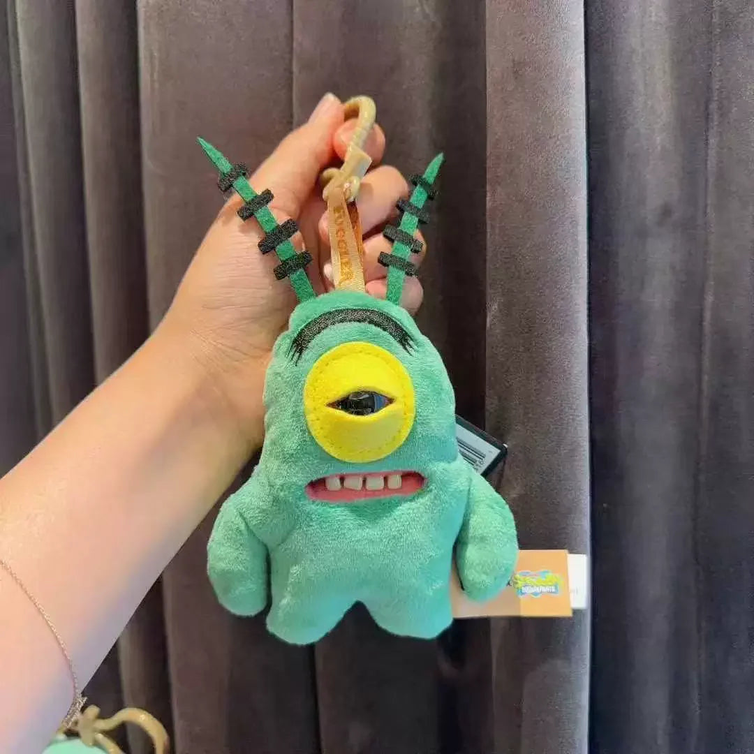 A hand holds a green monster plush keychain with spiky features and a yellow eye.