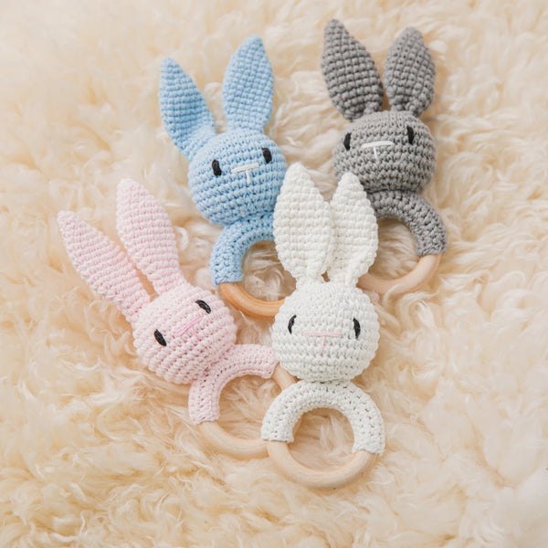 A set of handmade crochet bunny rattles in light blue, gray, and pink resting on a fluffy beige surface.
