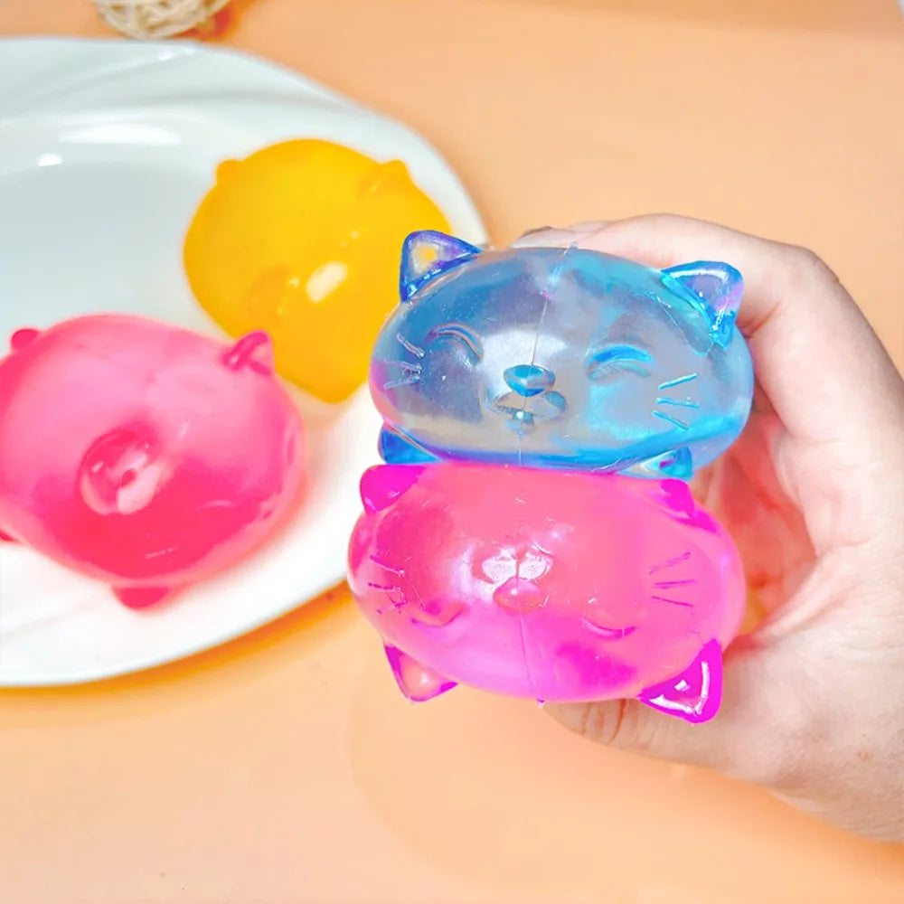 A hand holds a translucent blue cat squishy toy next to a pink one and a yellow one on a white plate.