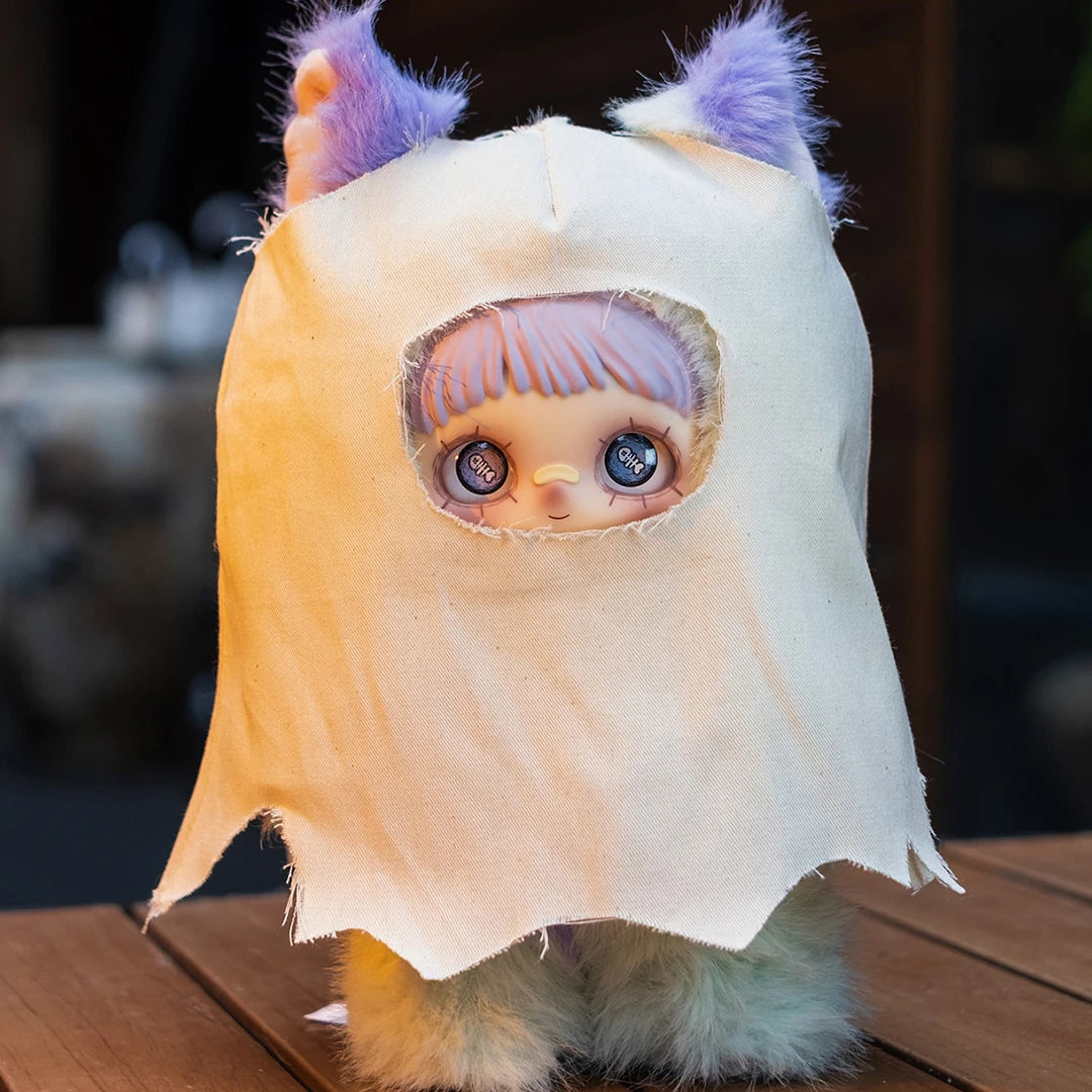 A light beige plush doll with purple hair and large blue eyes wearing a draped fabric hood.