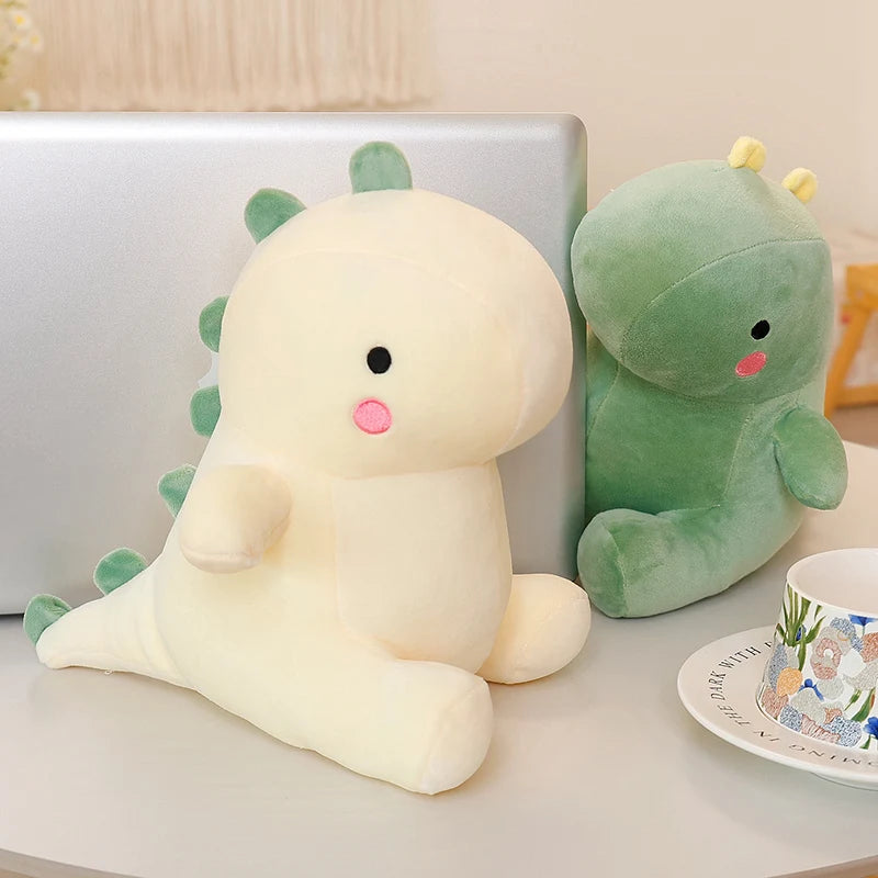A soft, pale yellow dinosaur plush toy resting on a white surface next to a silver laptop.
