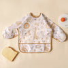 A white feeding bib with a light brown pattern of animals and food, shown flat on a beige surface.