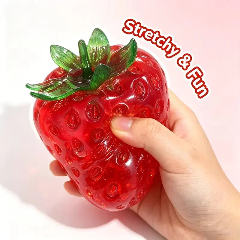 A hand holds a bright red strawberry squishy stress ball with green leaves, demonstrating its stretchy and fun texture.