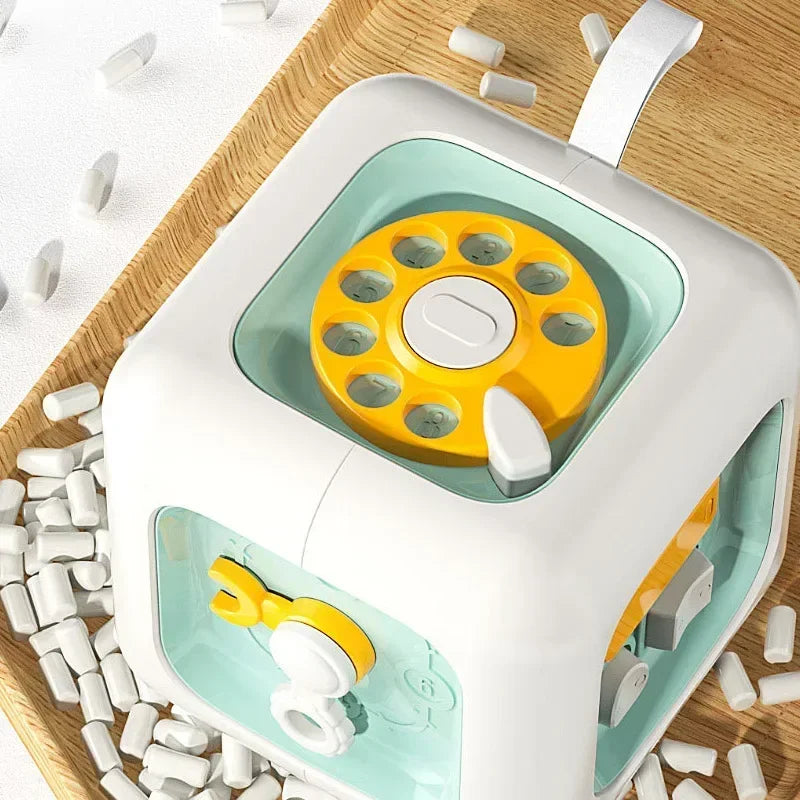 A white and teal Montessori busy cube toy with a yellow rotating disk and headphones is shown on a wooden surface.