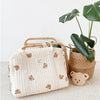 A cream quilted diaper bag with brown embroidery rests next to a potted tropical plant.