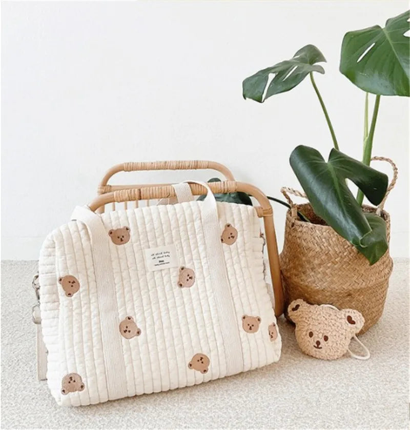 A cream quilted diaper bag with brown embroidery rests next to a potted tropical plant.
