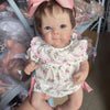 A lifelike 18-inch reborn baby doll with hand-painted details wearing a white floral dress and pink bow