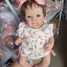 A lifelike 18-inch reborn baby doll with hand-painted details wearing a white floral dress and pink bow