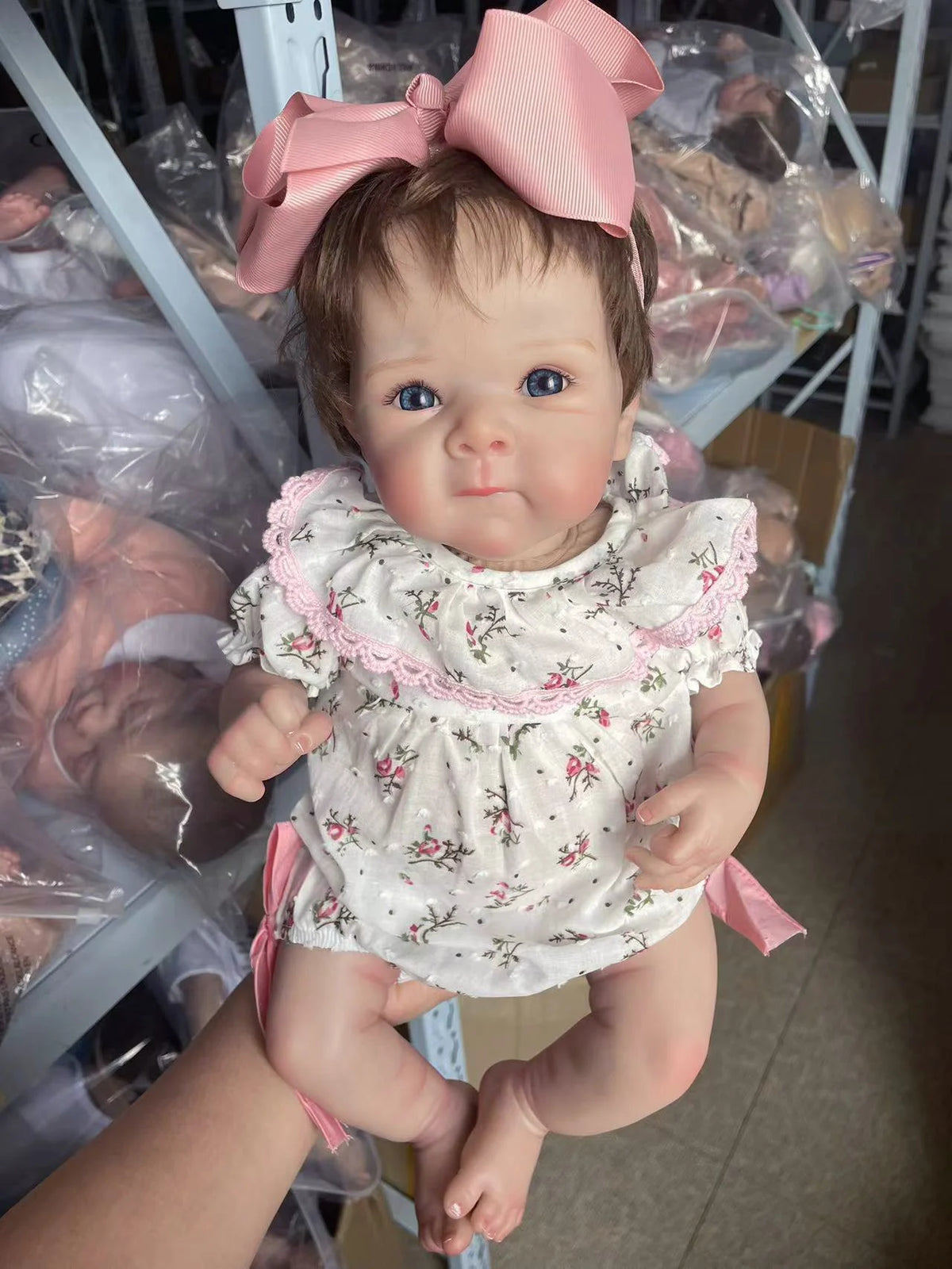 A lifelike 18-inch reborn baby doll with hand-painted details wearing a white floral dress and pink bow