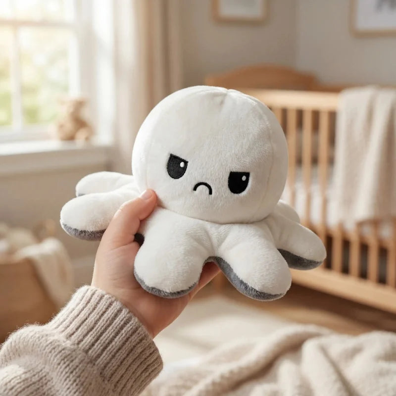 A hand is holding a white flip octopus plush toy with a grumpy expression in a bright indoor setting.