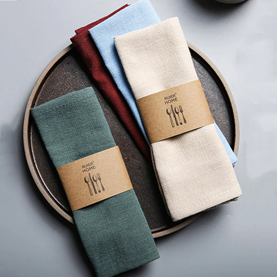 A set of four folded linen napkins in shades of blue, green, beige, and burgundy arranged on a wooden plate.