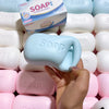 A hand holds a light blue soap ball squishy fidget toy surrounded by other white and pink soap products.