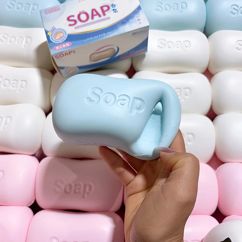 A hand holds a light blue soap ball squishy fidget toy surrounded by other white and pink soap products.