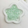 A white flower-shaped dish filled with green iridescent acrylic ice strips for nail art and DIY crafts.
