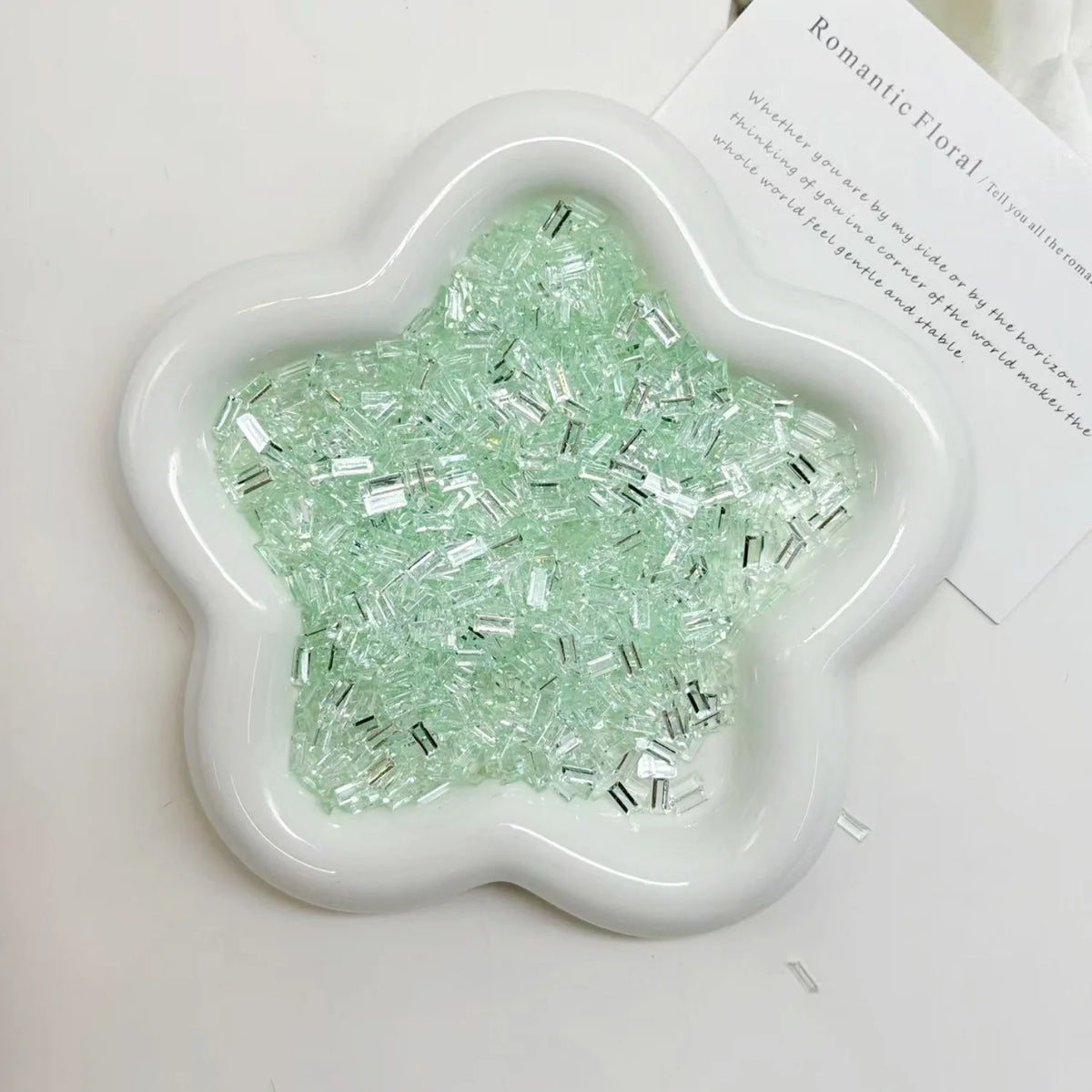 A white flower-shaped dish filled with green iridescent acrylic ice strips for nail art and DIY crafts.
