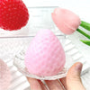 A pink strawberry squishy toy is shown resting in a clear glass dish, with a red strawberry visible nearby.