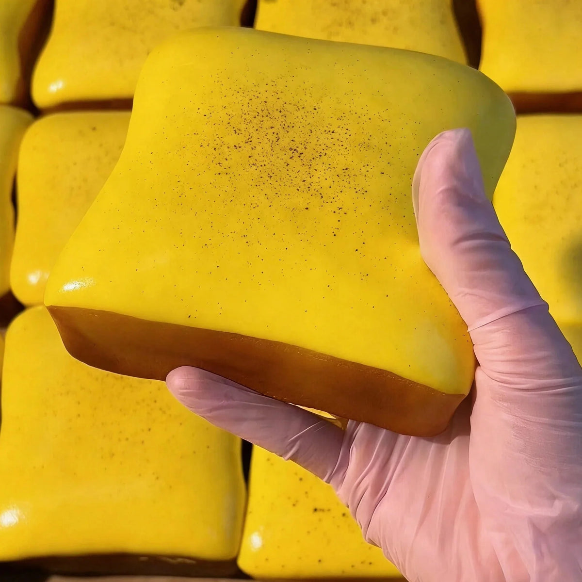 A hand in a pink glove holds a bright yellow, squishy cube toy.