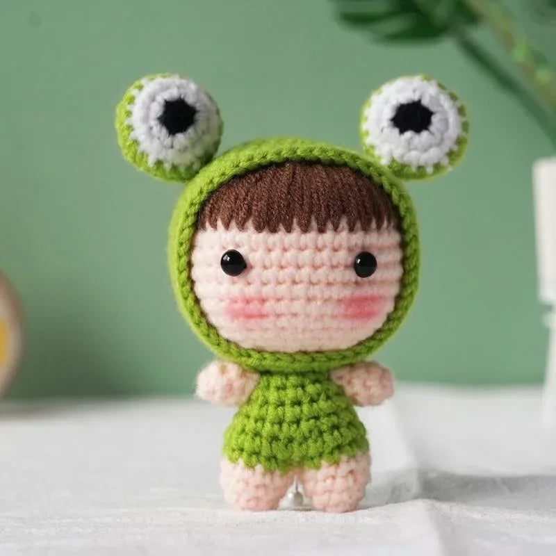 A handmade crochet doll kit featuring a green body, brown hair, and large white eyes with black pupils.