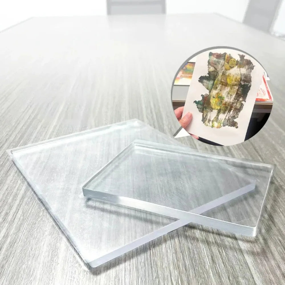 A clear silicone gel printing plate is shown on a wooden surface next to a small piece of art.