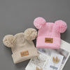 A pink knitted baby beanie hat with fluffy pom-poms and a beige version displayed on a white background.