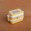 A miniature doll picnic lunch box in yellow and white with a floral design on the lid.