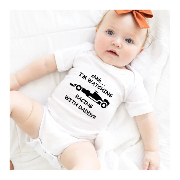 A baby wearing a white short-sleeved shirt with text that says I'm watching racing with daddy, lying on a white blanket.