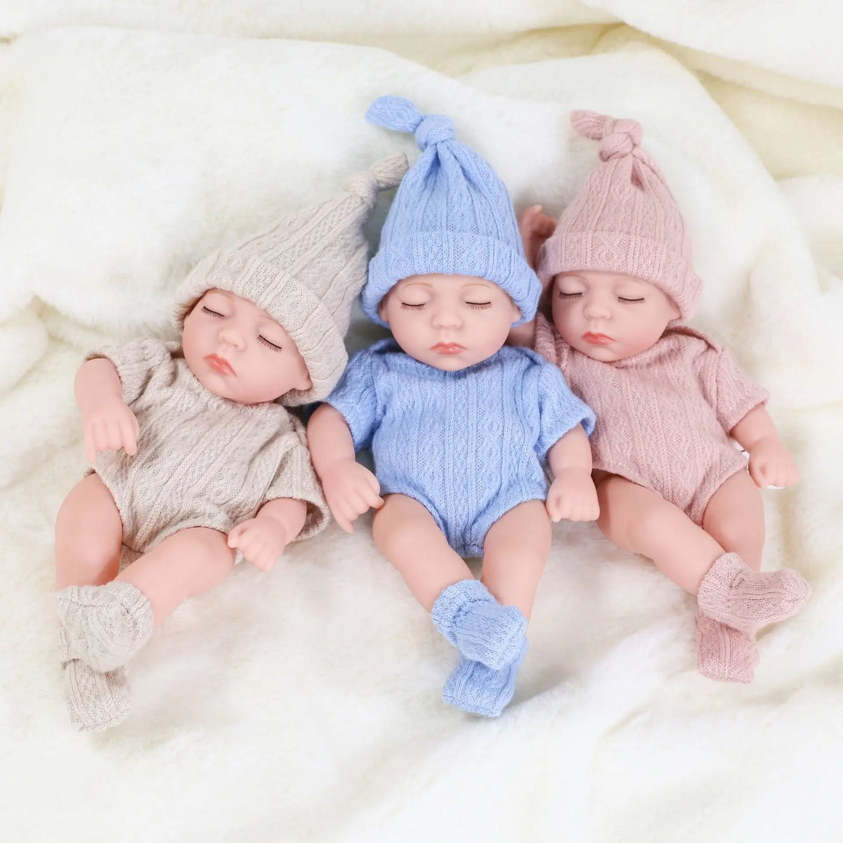 Three realistic soft vinyl baby dolls wearing knitted outfits lying on a white fluffy blanket