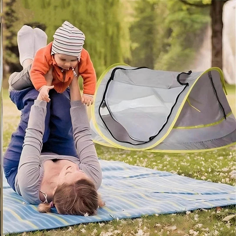 A baby is sitting in a gray instant pop-up tent with yellow trim outdoors on a blanket.