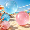 A hand holds a pink Mochi squishy stress relief toy on a sandy beach with a tennis court in the background.
