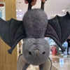A soft grey bat plush toy with large wings is being held up against a blurred indoor background.
